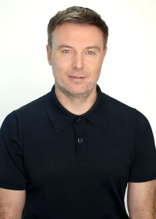 Man with short brown hair wearing a black polo shirt, posing against a plain light background.