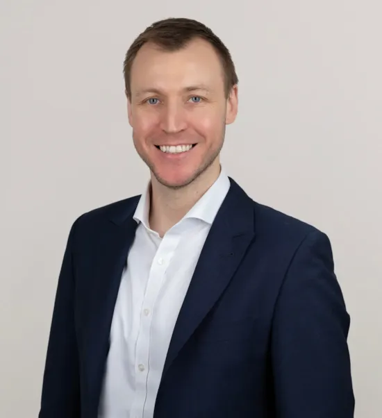 Smiling man in a navy blue suit and white shirt poses against a plain, light-colored background.