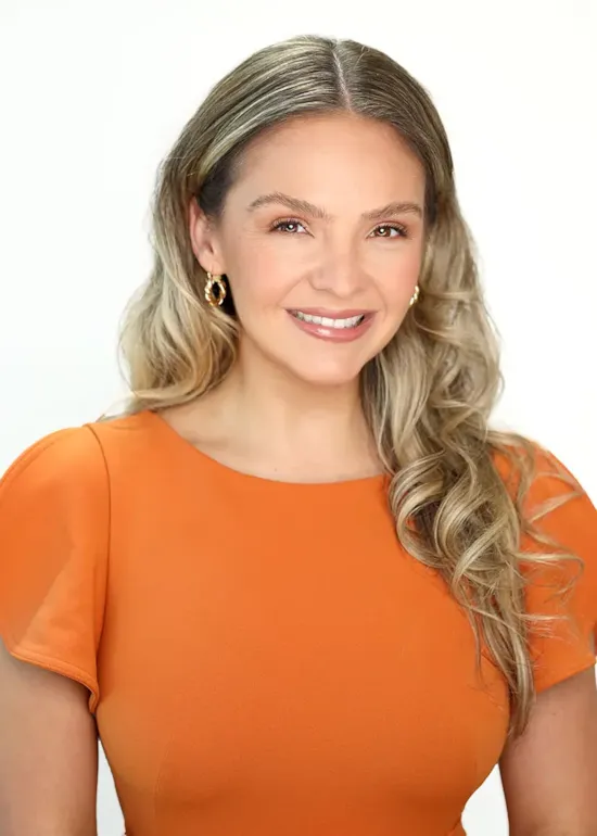 Smiling woman with long, wavy blonde hair wearing an orange top and gold hoop earrings on a white background.