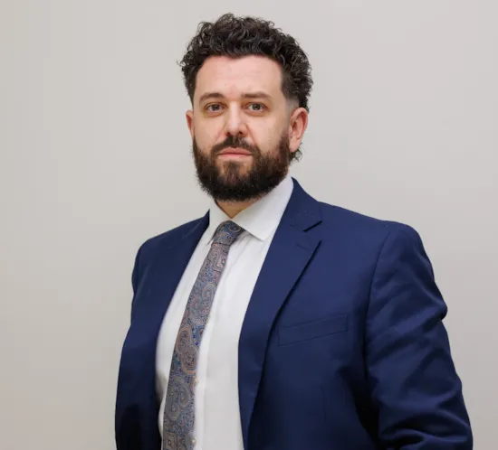 Man with shaggy/curly brown hair, dark brown beard, wearing a blue suit jacket and white shirt, smiling at the camera.