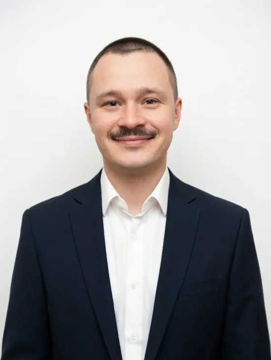 A man in a dark suit and white shirt stands smiling in front of a plain white wall.