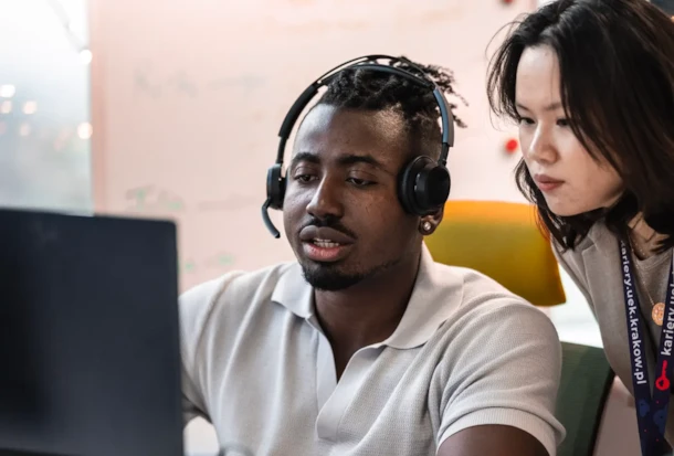 Two people looking at a computer screen; one is wearing a headset, and the other is leaning in to assist.