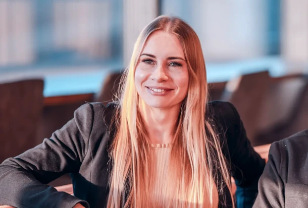 A young woman with long blonde hair smiles while sitting indoors, wearing a blazer and light-colored top.