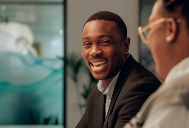 A man in a suit smiles while talking to another person in an office setting.
