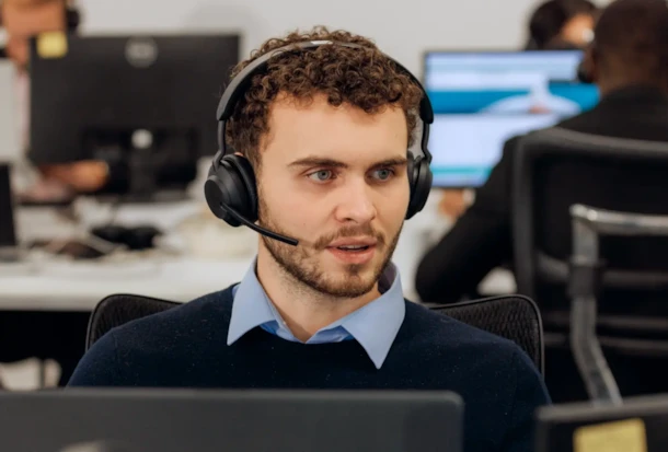A man wearing a headset works at a computer in an office with other people in the background.