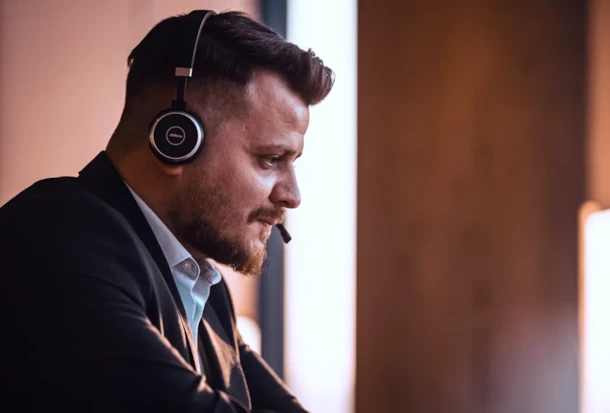 Man with beard wearing a headset and suit jacket, looking focused while working in an office environment.