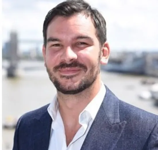 Man with dark hair and beard in a blue suit jacket, smiling outdoors by a river with cityscape in the background.